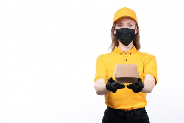 Un giovane corriere femminile di vista frontale in guanti neri uniformi gialli e maschera nera che tiene pacchetto dell'alimento