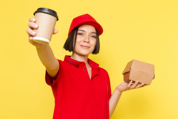 Un giovane corriere femminile di vista frontale in guanti neri uniformi rossi e spiritello malevolo che tiene pacchetto dell'alimento e tazza di caffè
