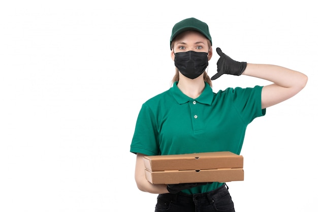 Un giovane corriere femminile di vista frontale in guanti neri uniformi verdi e maschera nera che tiene le scatole di consegna degli alimenti