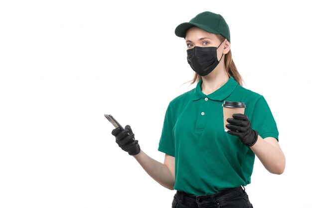 Un giovane corriere femminile di vista frontale in guanti neri uniformi verdi e maschera nera che tiene tazza di caffè e telefono