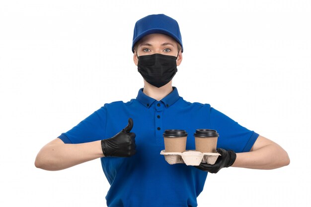 Un giovane corriere femminile di vista frontale in maschera nera uniforme blu e guanti che tengono le tazze di caffè