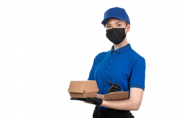 Un giovane corriere femminile di vista frontale in maschera nera uniforme blu e guanti che tengono pacchetto e blocco note di consegna dell'alimento