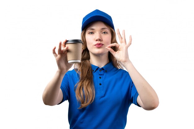Un giovane corriere femminile di vista frontale in tazza blu che tiene tazza di caffè della tenuta dello spiritello malevolo che mostra segno saporito su bianco