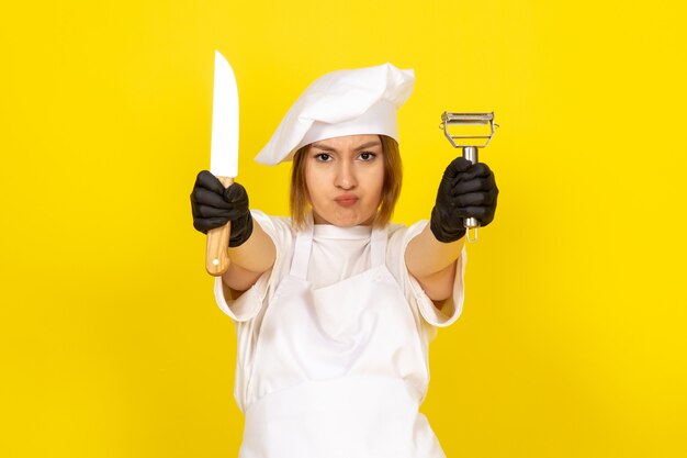 Un giovane cuoco femmina di vista frontale in abito bianco cuoco e berretto bianco in guanti neri tenendo coltello e pulitore di verdure sul giallo