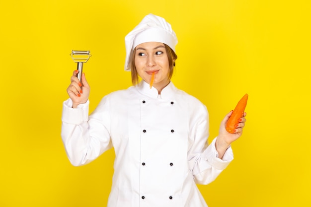 Un giovane cuoco femminile di vista frontale in vestito bianco del cuoco e protezione bianca che tiene e che pulisce carota arancio che sorride sul giallo