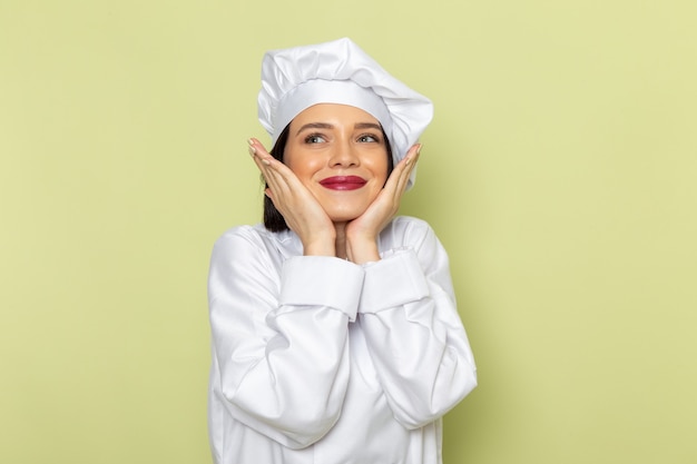 Un giovane cuoco femminile di vista frontale in vestito bianco e cappuccio del cuoco che posano con l'espressione felice sul colore di cucina dell'alimento del lavoro della signora della parete verde