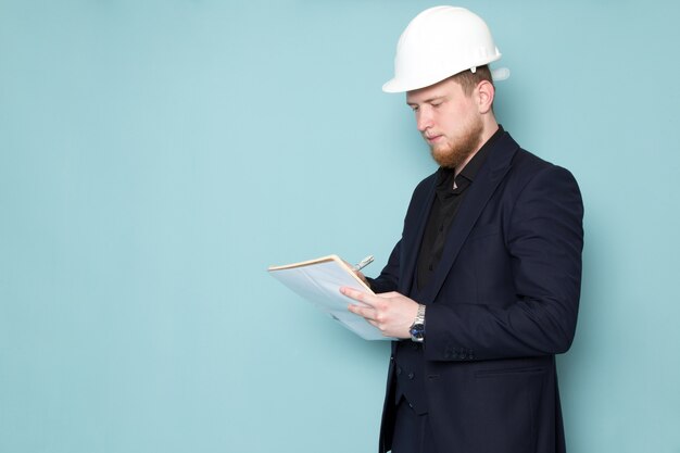 Un giovane maschio attraente di vista frontale con la barba in vestito moderno classico scuro nero nel casco bianco della costruzione che annota le note sullo spazio blu