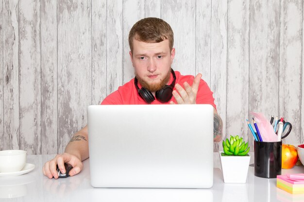 Un giovane maschio di vista frontale in camicia rosa in cuffie nere facendo uso della tavola grigia del computer portatile con lo spazio di gray della pianta del caffè delle penne