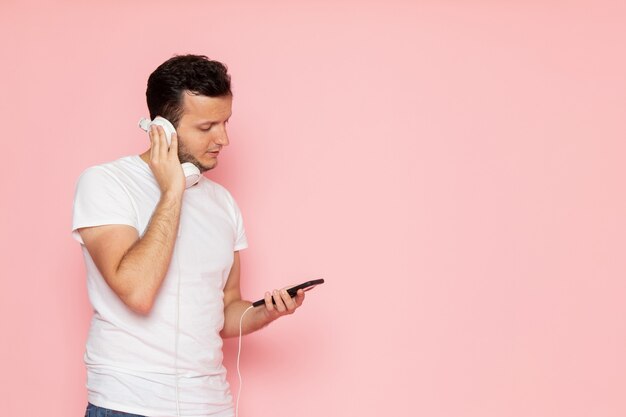 Un giovane maschio di vista frontale in maglietta bianca facendo uso degli auricolari del telefono sulla posa rosa di emozione di colore dello scrittorio