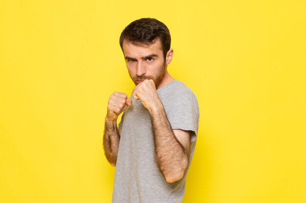 Un giovane maschio di vista frontale in maglietta grigia che posa con il supporto di boxe sul modello di colore di emozione di espressione dell'uomo della parete gialla