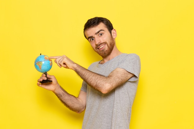 Un giovane maschio di vista frontale in maglietta grigia che tiene piccolo globo con il sorriso sui vestiti gialli di emozione del modello di colore dell'uomo della parete