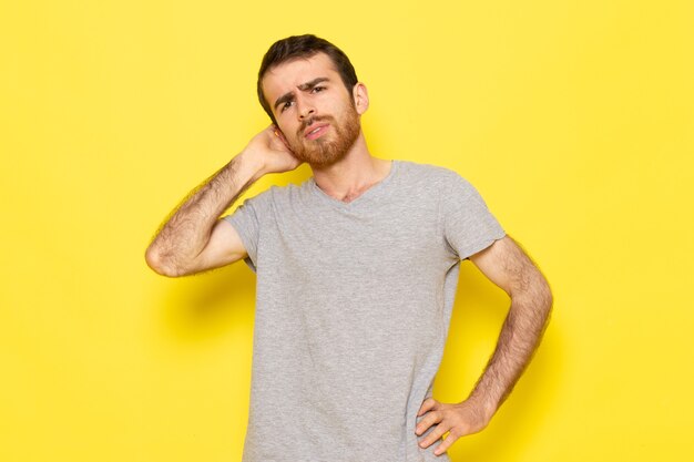 Un giovane maschio di vista frontale in maglietta grigia con l'espressione di pensiero sul modello di colore di emozione di espressione dell'uomo della parete gialla