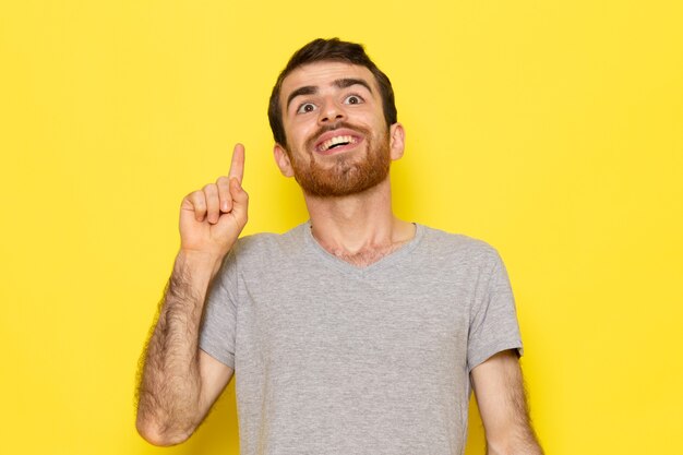 Un giovane maschio di vista frontale in maglietta grigia con l'espressione divertente sul modello di colore di emozione di espressione dell'uomo della parete gialla