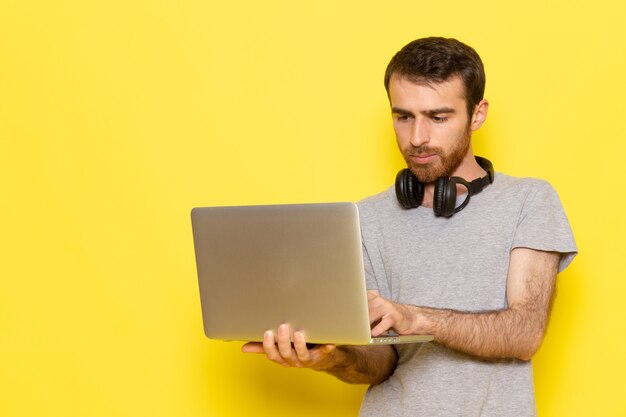 Un giovane maschio di vista frontale in maglietta grigia facendo uso del computer portatile sul modello di colore di colore di emozione di espressione dell'uomo della parete gialla