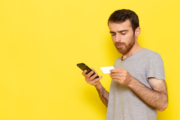 Un giovane maschio di vista frontale in maglietta grigia usando il suo telefono sui vestiti gialli di emozione del modello di colore dell'uomo della parete