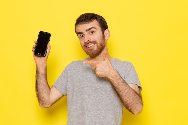 Un giovane maschio di vista frontale in t-shirt grigia che tiene smartphone con il sorriso sul modello di colore di emozione di espressione dell'uomo della parete gialla