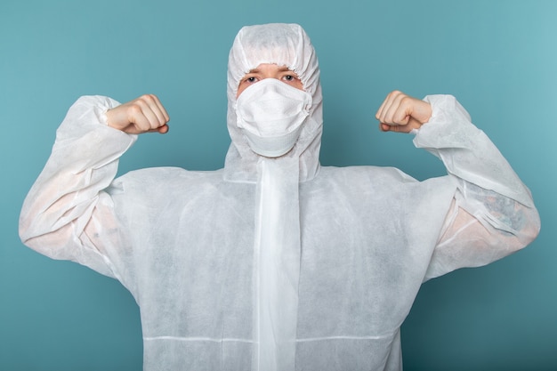 Un giovane maschio di vista frontale in vestito speciale bianco che porta maschera protettiva sterile che flette sul colore blu dell'attrezzatura speciale del pericolo del vestito dell'uomo della parete