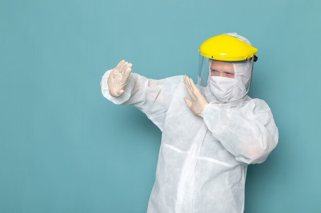 Un giovane maschio di vista frontale in vestito speciale bianco e casco speciale giallo che mantiene la distanza sul colore blu dell'attrezzatura speciale del pericolo del vestito dell'uomo della parete