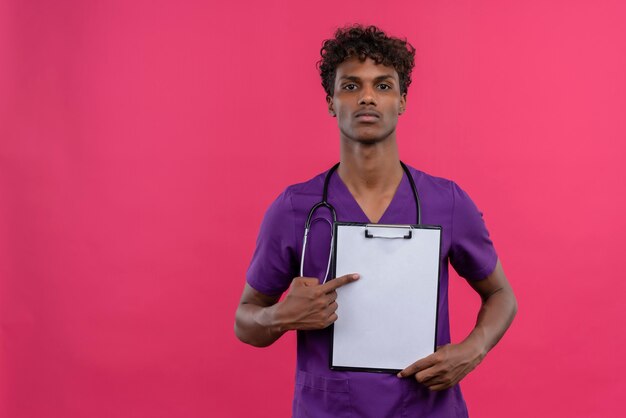 Un giovane medico dalla carnagione scura bello con capelli ricci che indossa l'uniforme viola con lo stetoscopio che indica con il dito indice negli appunti con un foglio di carta bianco