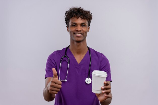 Un giovane medico dalla carnagione scura bello con capelli ricci che indossa l'uniforme viola con lo stetoscopio che tiene tazza di caffè di carta con i pollici in su