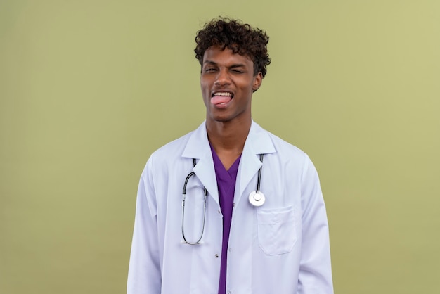 Un giovane medico dalla carnagione scura bello con capelli ricci che porta camice bianco con lo stetoscopio che mostra la sua lingua su uno spazio verde