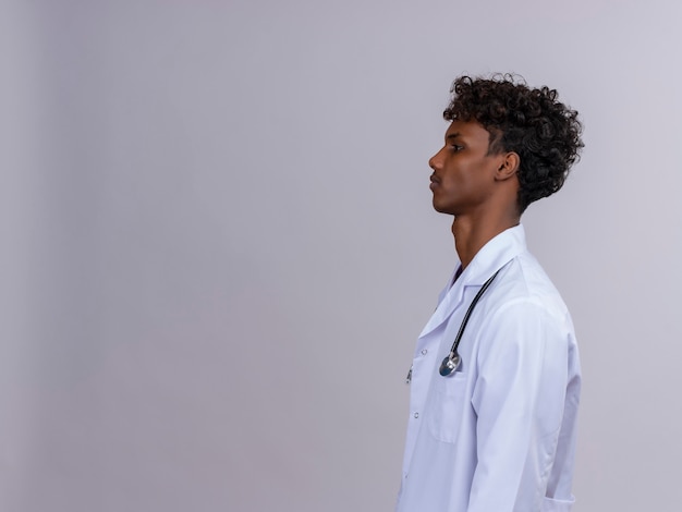 Un giovane medico dalla carnagione scura bello con capelli ricci che porta camice bianco con lo stetoscopio che osserva da parte