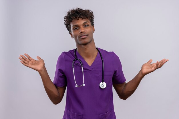 Un giovane medico dalla carnagione scura bello con capelli ricci che porta l'uniforme viola con lo stetoscopio che apre le mani con espressione confusa