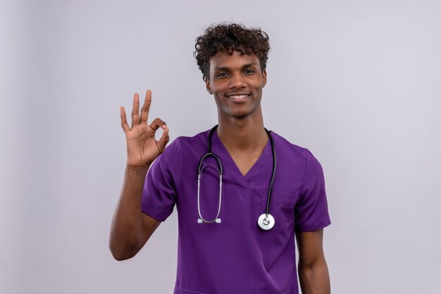 Un giovane medico dalla carnagione scura bello felice con capelli ricci che porta l'uniforme viola con lo stetoscopio che mostra segno giusto con le mani