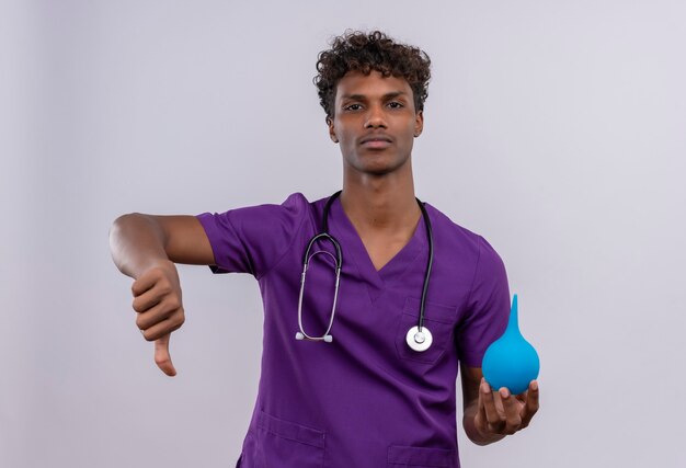 Un giovane medico dalla carnagione scura bello sconvolto con capelli ricci che indossa l'uniforme viola con lo stetoscopio che mostra i pollici giù mentre tiene un clistere