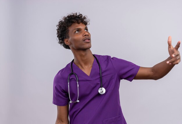 Un giovane medico dalla carnagione scura bello sicuro con capelli ricci che porta l'uniforme viola con lo stetoscopio che guarda verso l'alto mentre solleva le mani