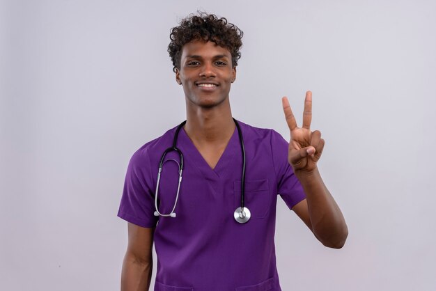 Un giovane medico dalla carnagione scura bello soddisfatto con capelli ricci che porta l'uniforme viola con lo stetoscopio che mostra il numero due con le dita