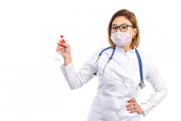 Un giovane medico femminile di vista frontale in vestito medico bianco con lo stetoscopio che indossa lo spruzzo bianco della tenuta della maschera protettiva sul bianco