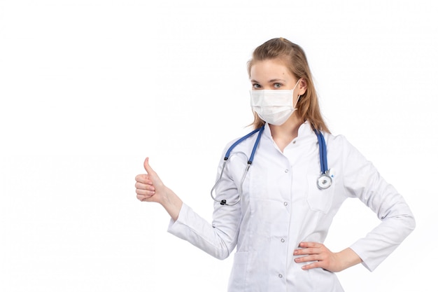 Un giovane medico femminile di vista frontale in vestito medico bianco con lo stetoscopio che indossa maschera protettiva bianca sul bianco
