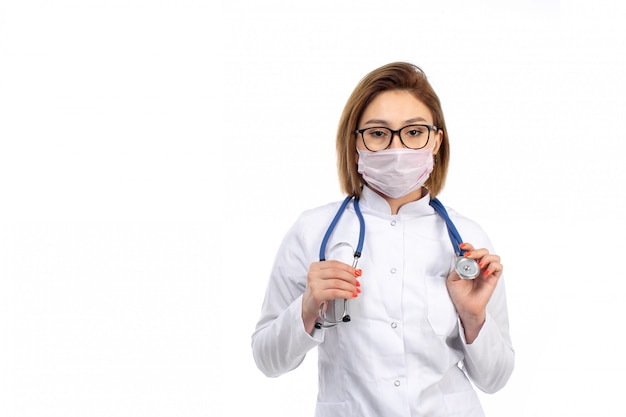 Un giovane medico femminile di vista frontale in vestito medico bianco con lo stetoscopio che indossa maschera protettiva bianca sul bianco