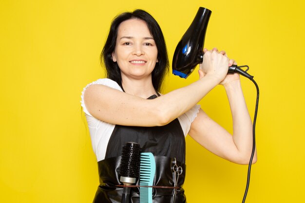 Un giovane parrucchiere femminile di vista frontale in mantello nero della maglietta bianca che tiene sorridere nero del fon