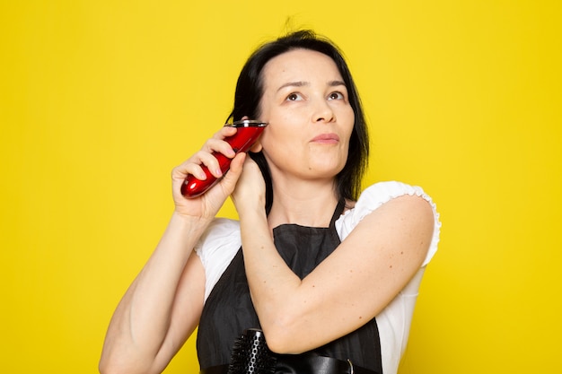 Un giovane parrucchiere femminile di vista frontale in mantello nero della maglietta bianca facendo uso dei capelli meccanici dell'onda della macchina dei capelli