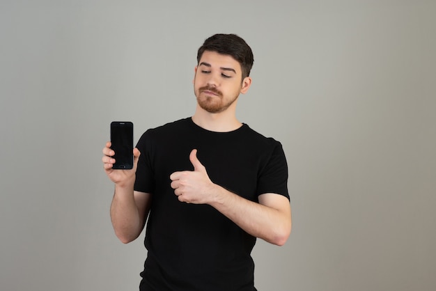 Un giovane ragazzo fiducioso in possesso di un telefono e gesticolando pollici in su su un grigio.