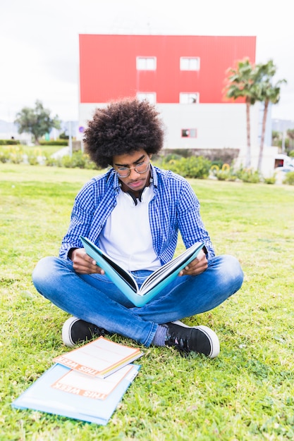 Un giovane studente maschio afroamericano che si siede sul prato che legge il libro