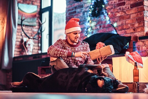 Un giovane uomo alla moda che tiene una scatola regalo mentre è seduto con il suo simpatico cane in un soggiorno decorato a Natale.