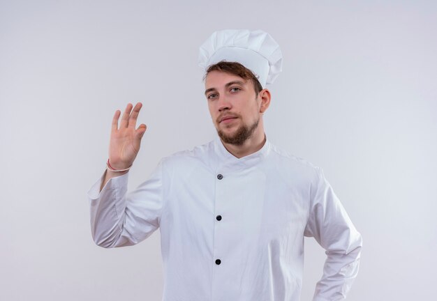 Un giovane uomo barbuto bello fiducioso del cuoco unico che indossa l'uniforme bianca del fornello e il cappello che mostra il gesto giusto saporito con le dita su una parete bianca