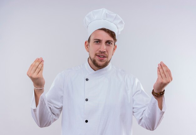 Un giovane uomo barbuto positivo del cuoco unico che indossa l'uniforme bianca del fornello e il cappello che tengono insieme le dita insieme il gesto della mano mentre osserva su una parete bianca
