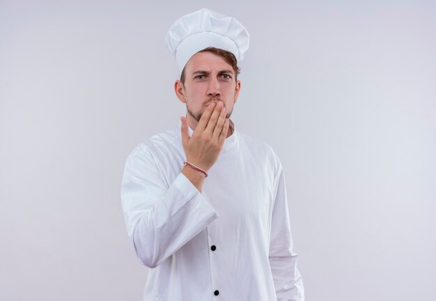 Un giovane uomo barbuto sorpreso del cuoco unico che porta l'uniforme e il cappello bianchi del fornello che tengono la mano sulla bocca mentre osservava su un muro bianco