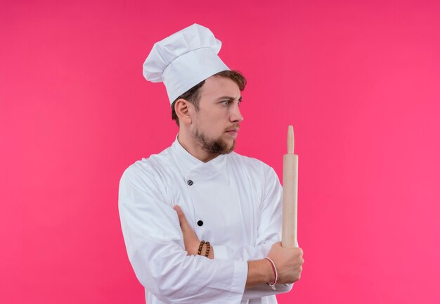 Un giovane uomo barbuto sorpreso del cuoco unico in uniforme bianca che tiene il mattarello mentre guarda il lato su una parete rosa