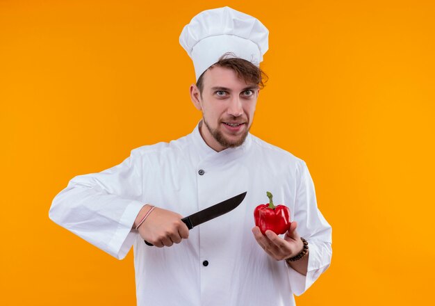 Un giovane uomo barbuto sorridente del cuoco unico in uniforme bianca che taglia il peperone rosso con il coltello mentre osserva su una parete arancione