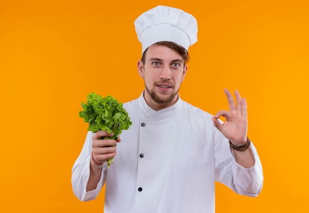 Un giovane uomo barbuto sorridente del cuoco unico in uniforme bianca che tiene la lattuga verde della foglia mentre osserva su una parete arancione