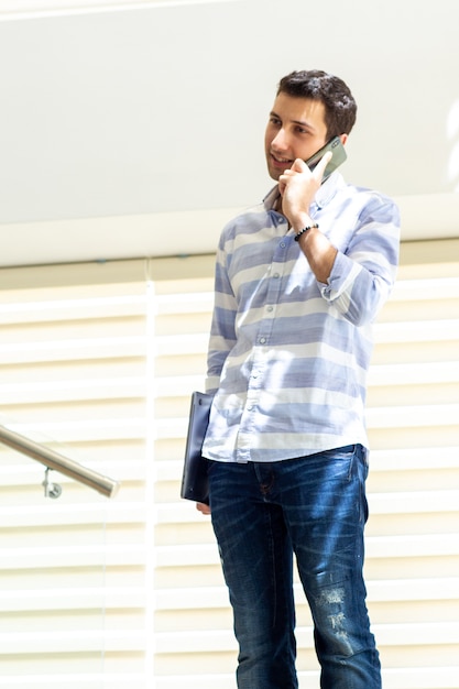 Un giovane uomo bello di vista frontale in camicia a strisce che parla e che discute le questioni di lavoro sul telefono durante la costruzione di attività di lavoro di giorno