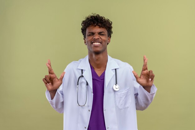 Un giovane uomo dalla carnagione scura bello con capelli ricci che porta camice bianco con lo stetoscopio che mostra le dita piegate su uno spazio verde