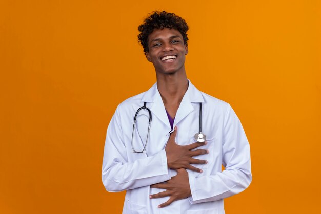 Un giovane uomo dalla carnagione scura bello con capelli ricci che porta camice bianco con lo stetoscopio che sorride e che tiene le mani