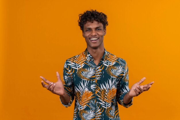 Un giovane uomo dalla carnagione scura bello con capelli ricci in camicia stampata foglie aprendo le mani e sorridendo