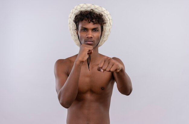 Un giovane uomo dalla carnagione scura bello scontento con i capelli ricci che porta il cappello del sole che mostra i movimenti di boxe mentre